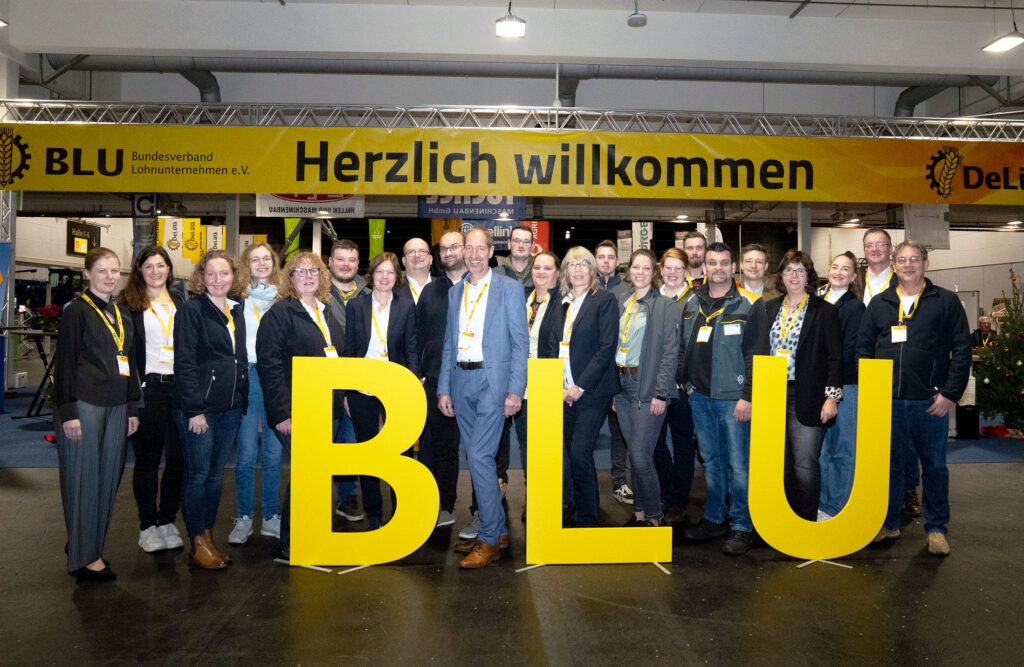 Eine Gruppe von Menschen steht lächelnd hinter großen gelben Buchstaben mit der Aufschrift BLU bei einer Indoor-Veranstaltung.
