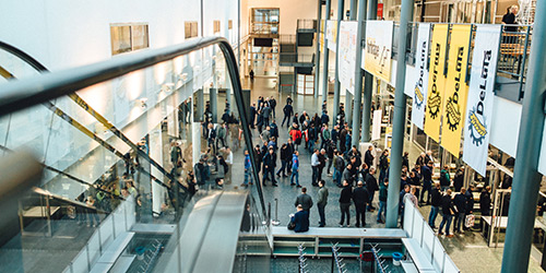 Eine große Gruppe von Menschen versammelt sich in einem modernen überdachten Atrium, in dem Transparente und eine Rolltreppe zu sehen sind.